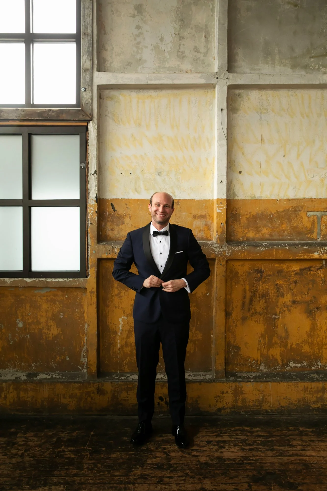 A man in a formal tuxedo with a bow tie standing indoors against a textured, weathered wall with large windows.