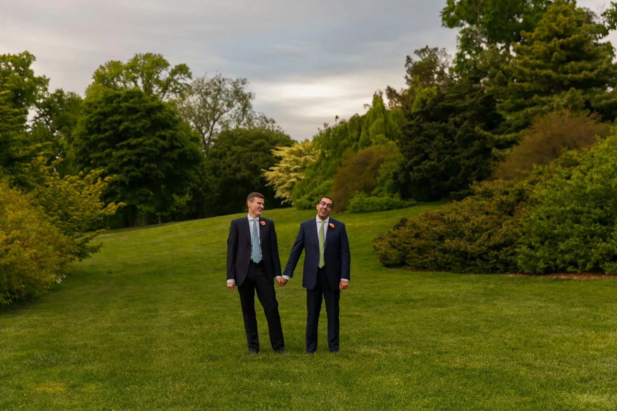 Two men in suits holding hands and smiling in a park with lush green trees and grass, under a cloudy sky.