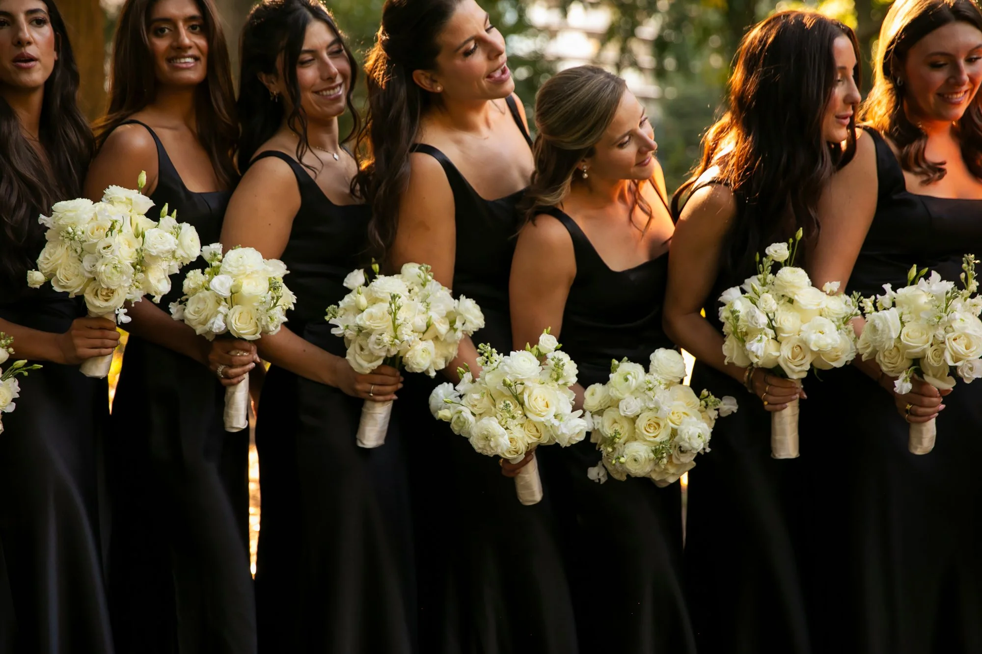 A group of bridesmaids dressed in black holding white bouquets, standing outdoors during sunset.