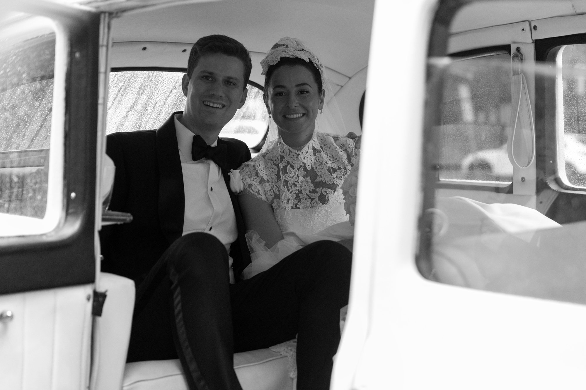 Black and white photo of a newlywed couple sitting inside a vintage car, smiling at the camera, with rain on the window.