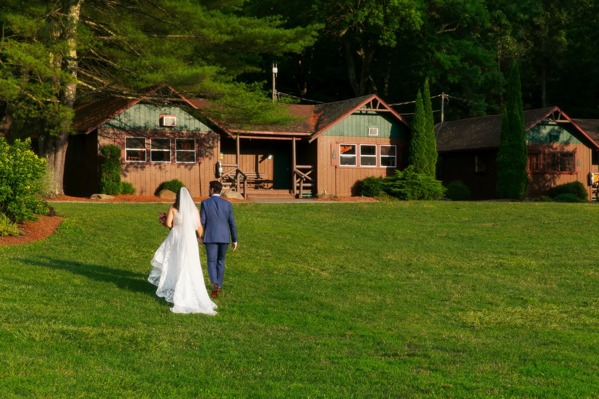 A bride in a white wedding dress and veil walking with a groom in a blue suit across a grassy lawn towards brown cabins with trees in the background.
