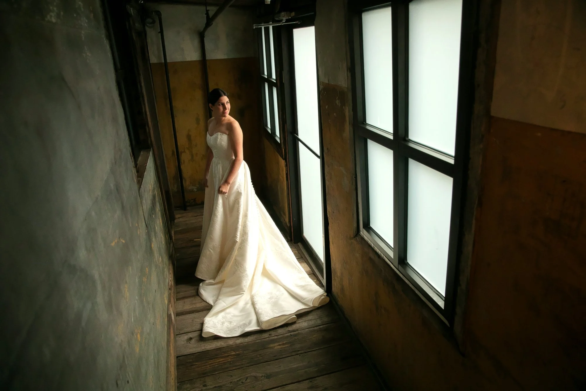 A woman in a white wedding dress standing in a narrow, rustic hallway with wooden floors and frosted windows.