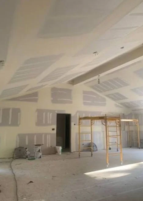 Interior of a room under construction with drywall and ceiling panels installed, scaffolding in the middle, and construction materials on the floor.