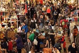 Busy indoor flea market with various vendors and shoppers browsing through tables of clothing, accessories, and collectibles.