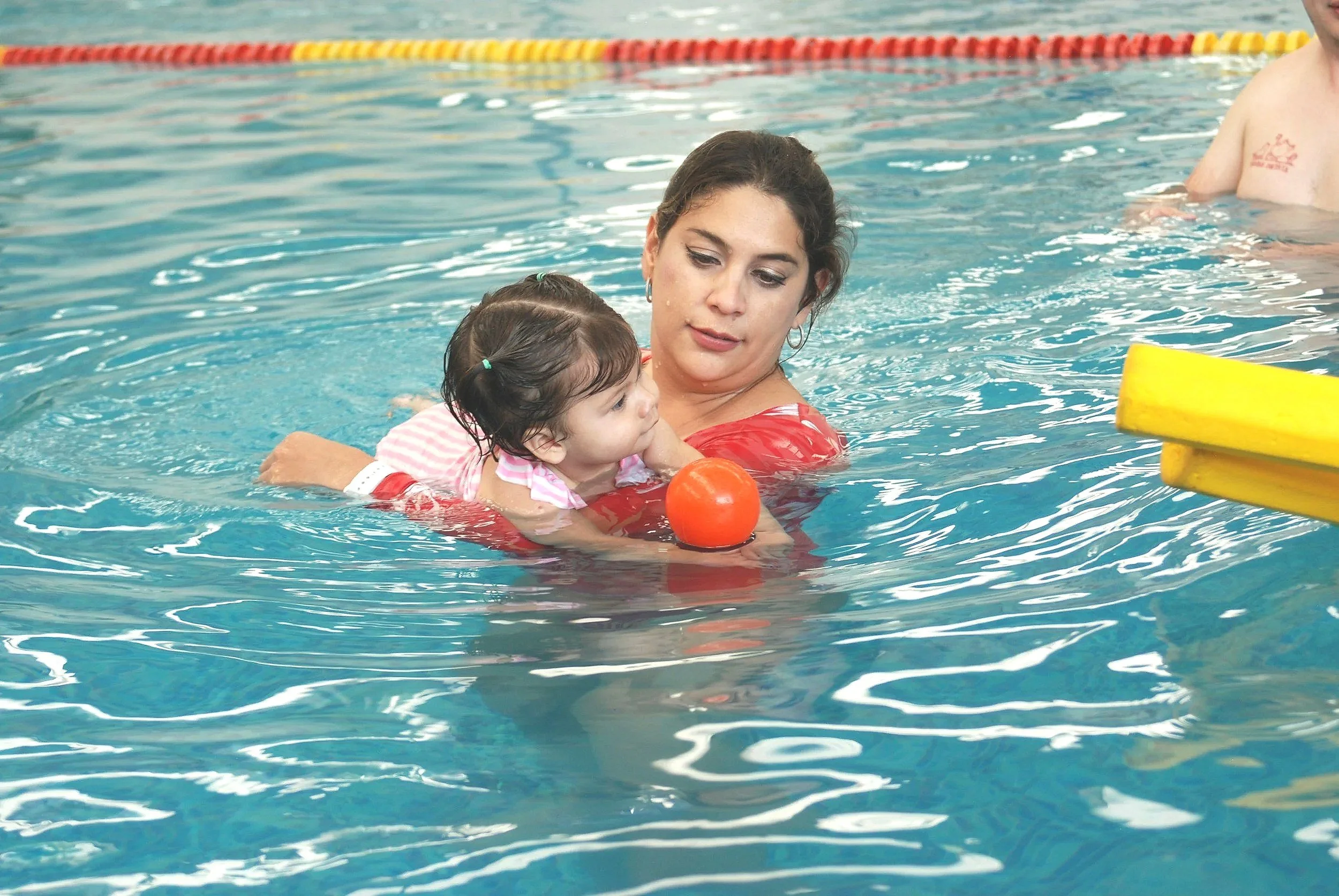 A woman teaching a young girl to swim in a swimming pool, with a red swimming float and an orange ball.
