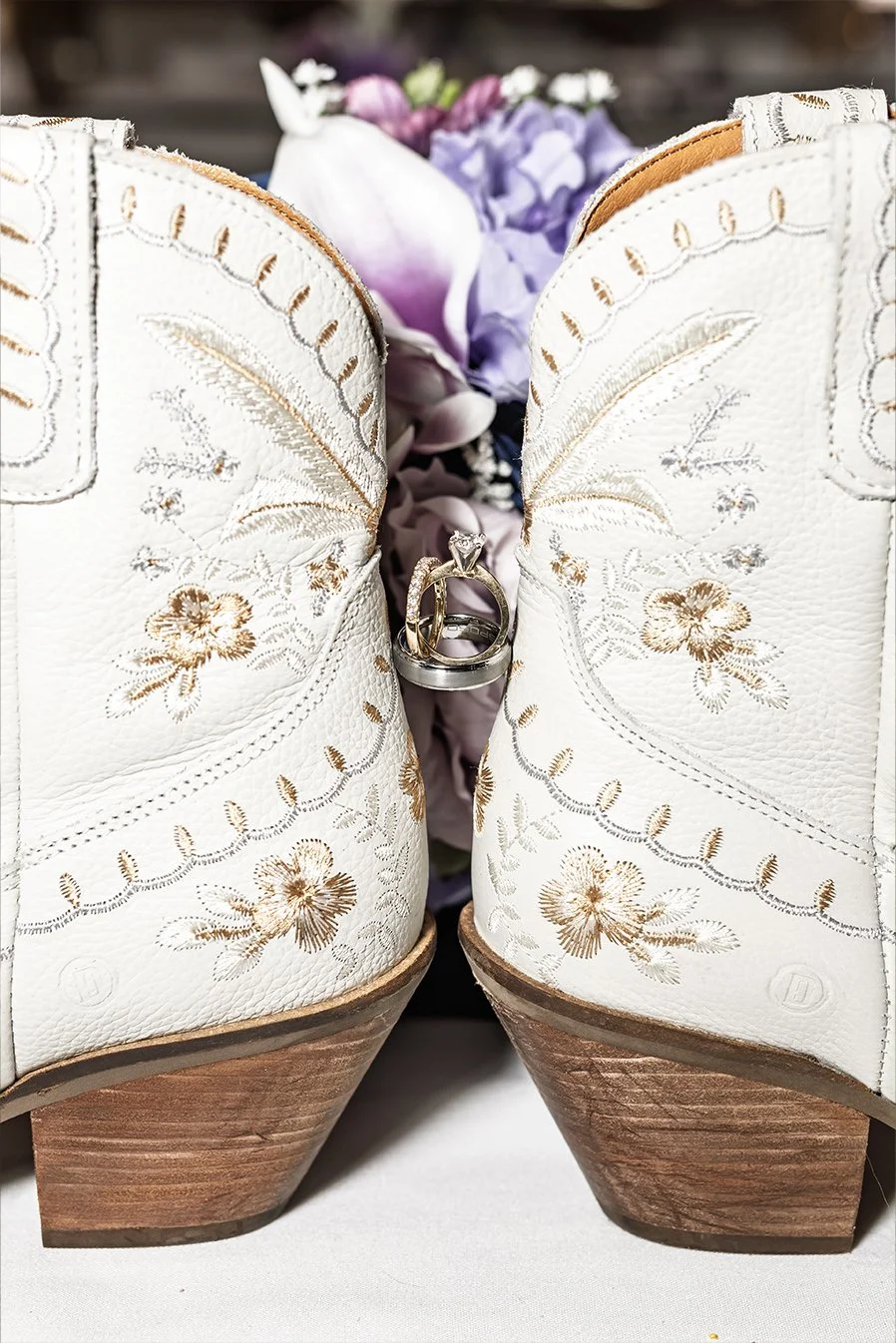 Pair of white cowboy boots with floral embroidery, wedding rings hanging between the boots, and purple flowers in the background.