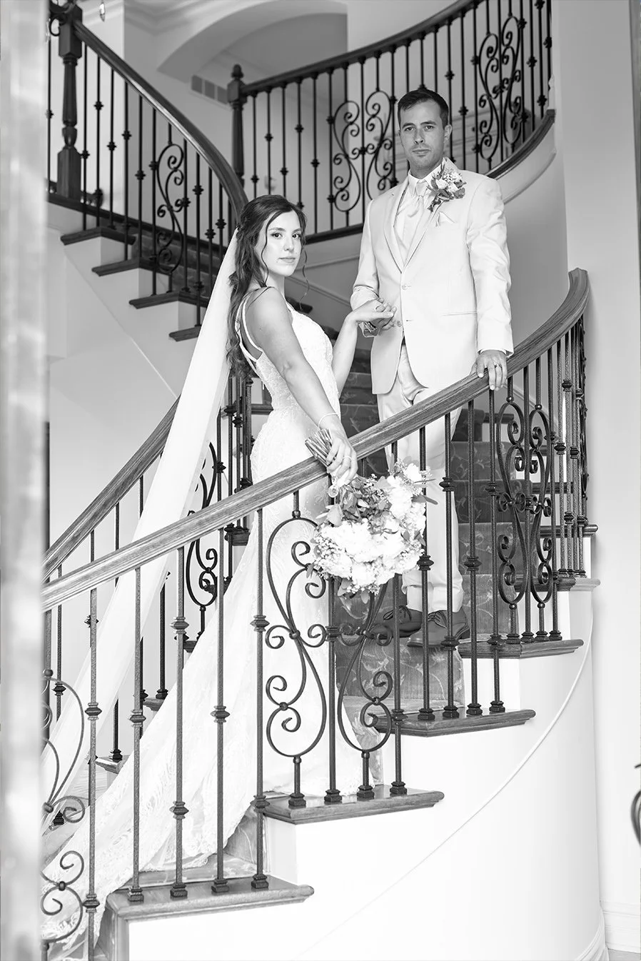 Black and white photo of a bride and groom on a staircase. The bride holds a bouquet, wearing a wedding dress and veil. The groom is in a light-colored suit with a boutonniere. They hold hands on the staircase railing.