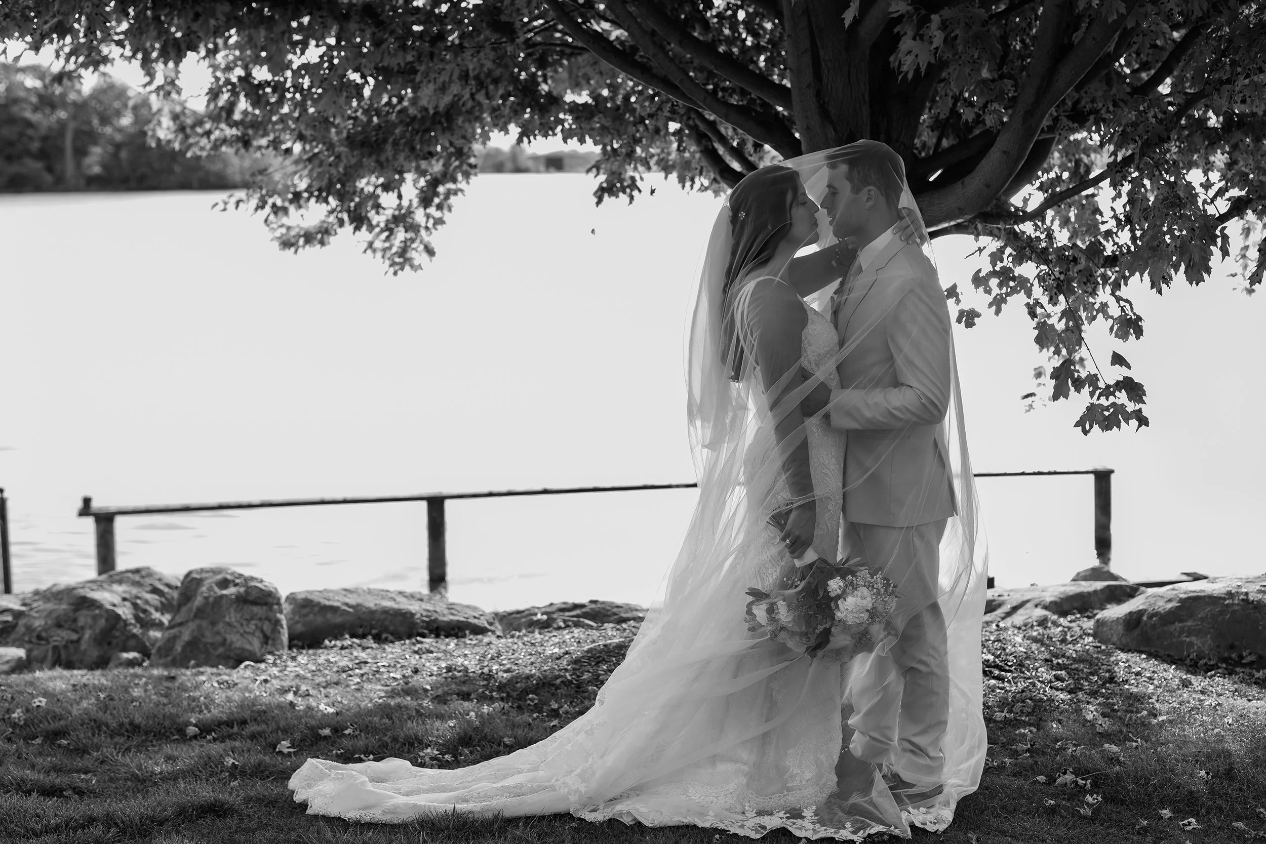 Black and white photo of a newlywed couple standing under a tree by a lake, with the bride holding a bouquet and both leaning in close, sharing a moment.
