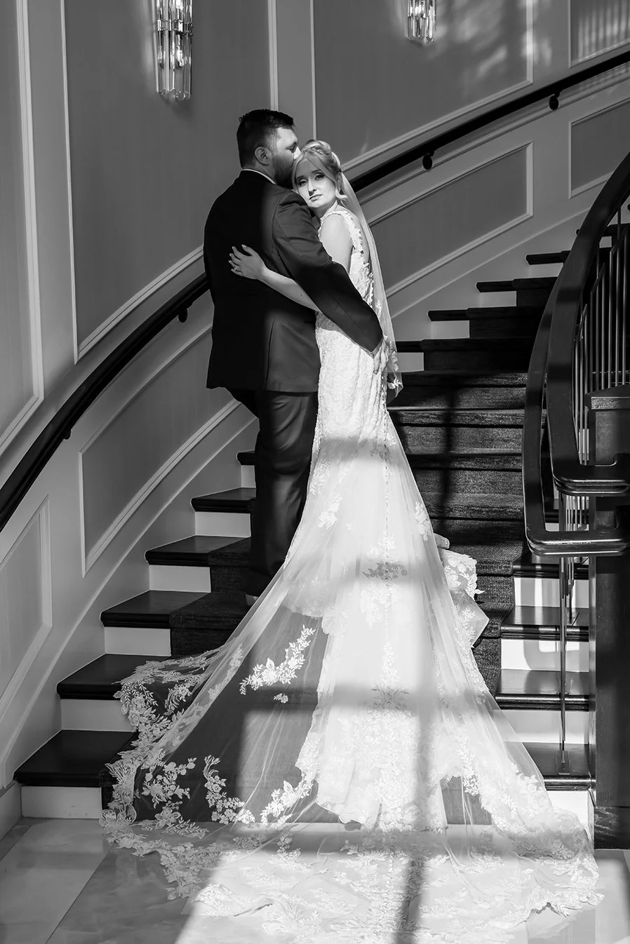 A black and white photo of a bride and groom hugging on a staircase during their wedding, with ornate wall lamps and detailed woodwork.