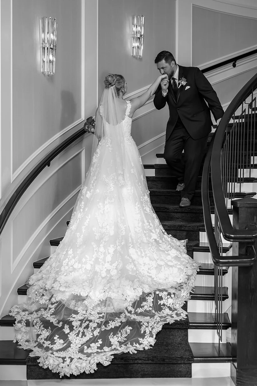A bride in a long, lace wedding dress is on a staircase, and a groom in a suit is kissing her hand.