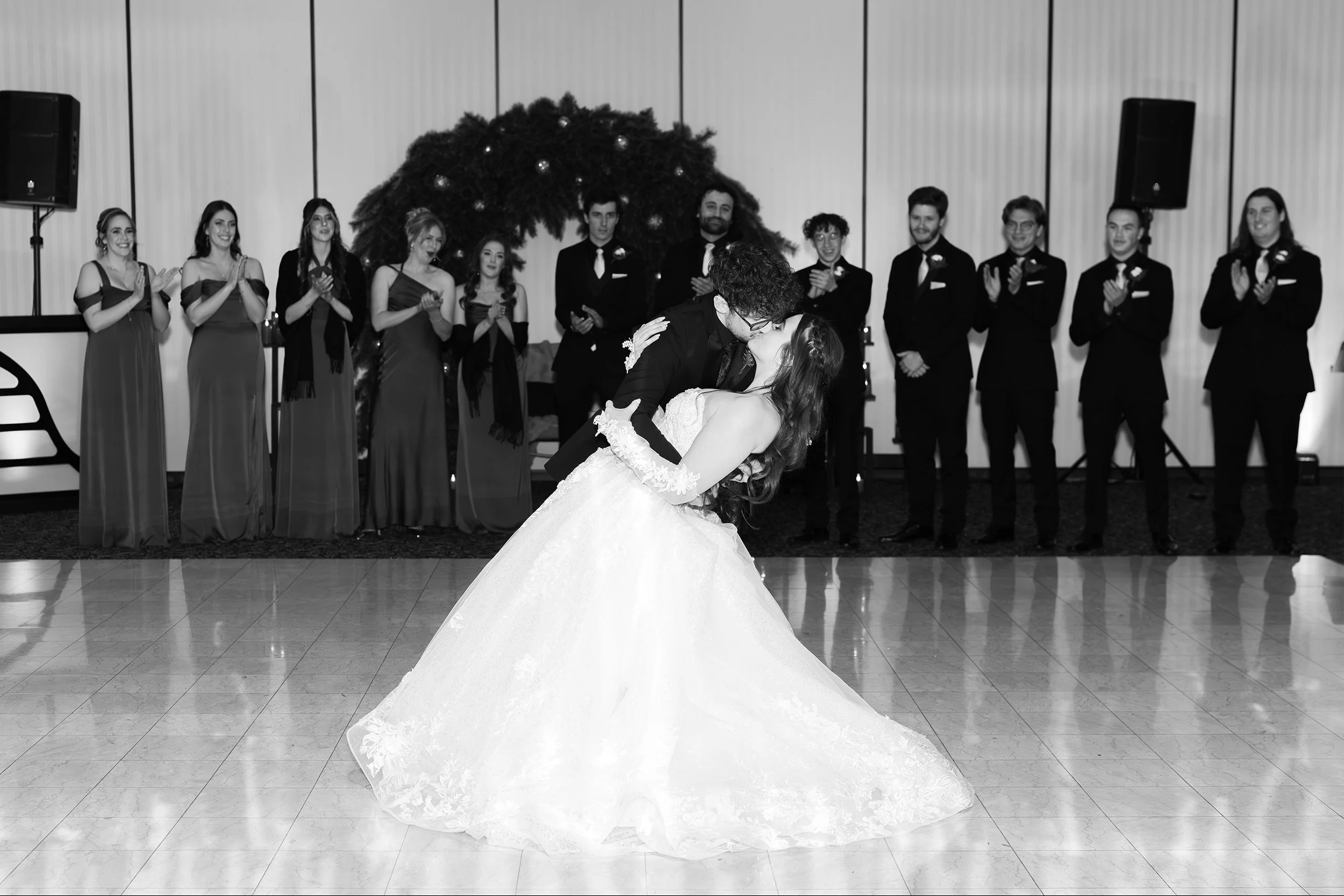 A black-and-white photo of a bride and groom sharing a first dance at their wedding reception, with a group of bridesmaids and groomsmen standing and clapping in the background.