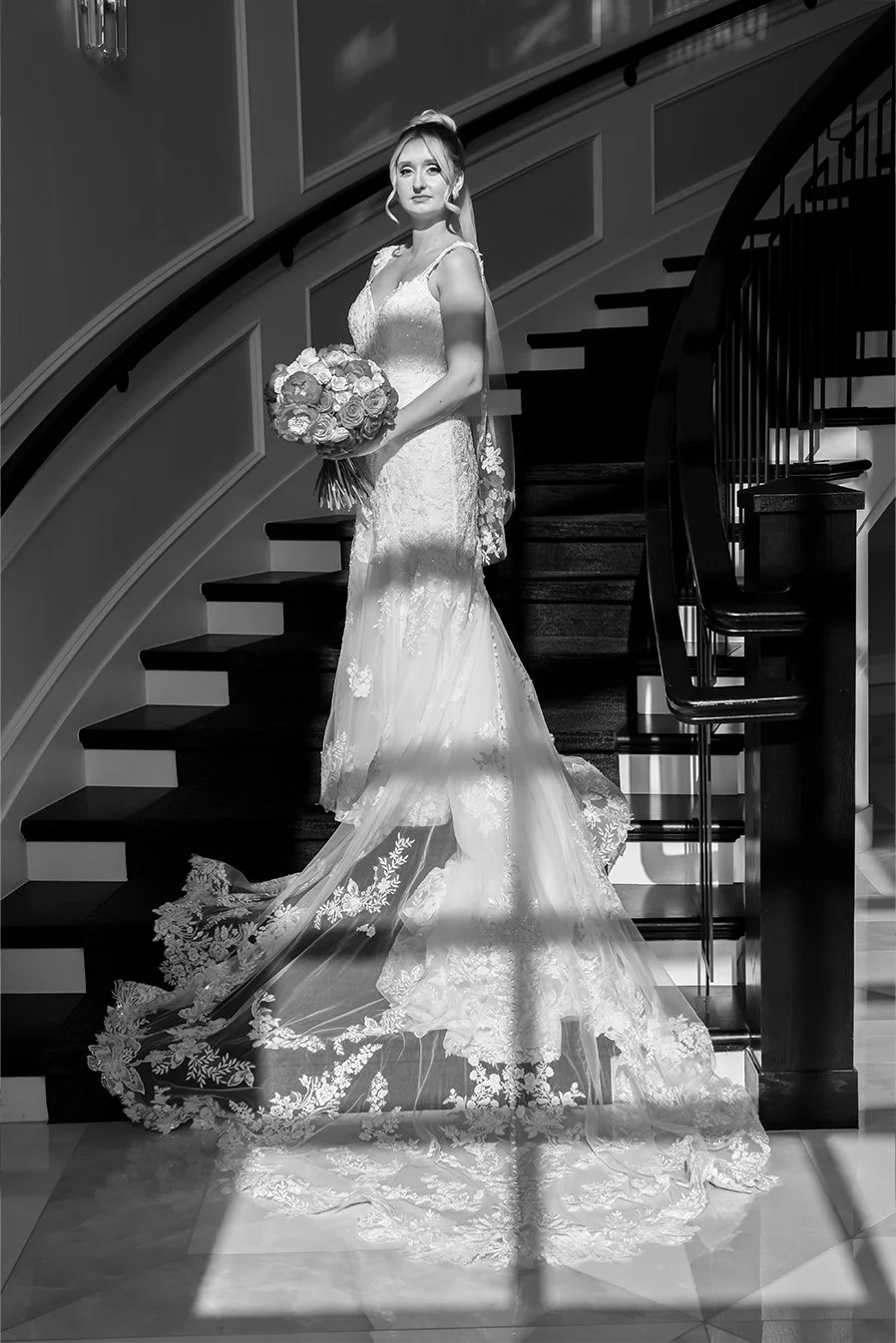 A bride in a wedding gown holding a bouquet of roses, standing on a staircase with carved wooden railings. Black and white editorial wedding photo