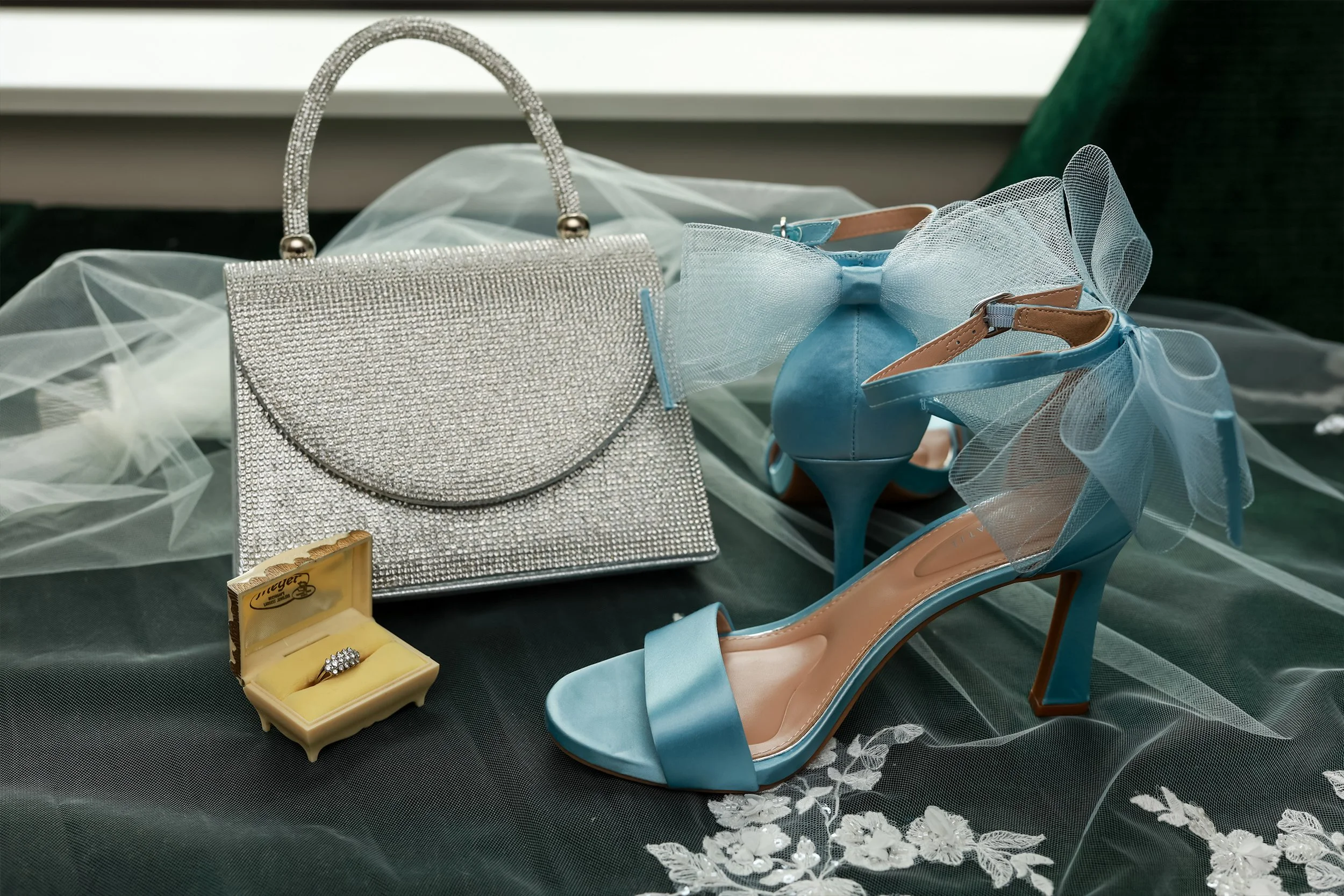 A pair of light blue high-heeled shoes with decorative tulle ribbons, a silver sparkly clutch, a small ring in a yellow jewelry box, and a light blue bow accessory, all arranged on a dark fabric with a sheer overlay. wedding details flat lay