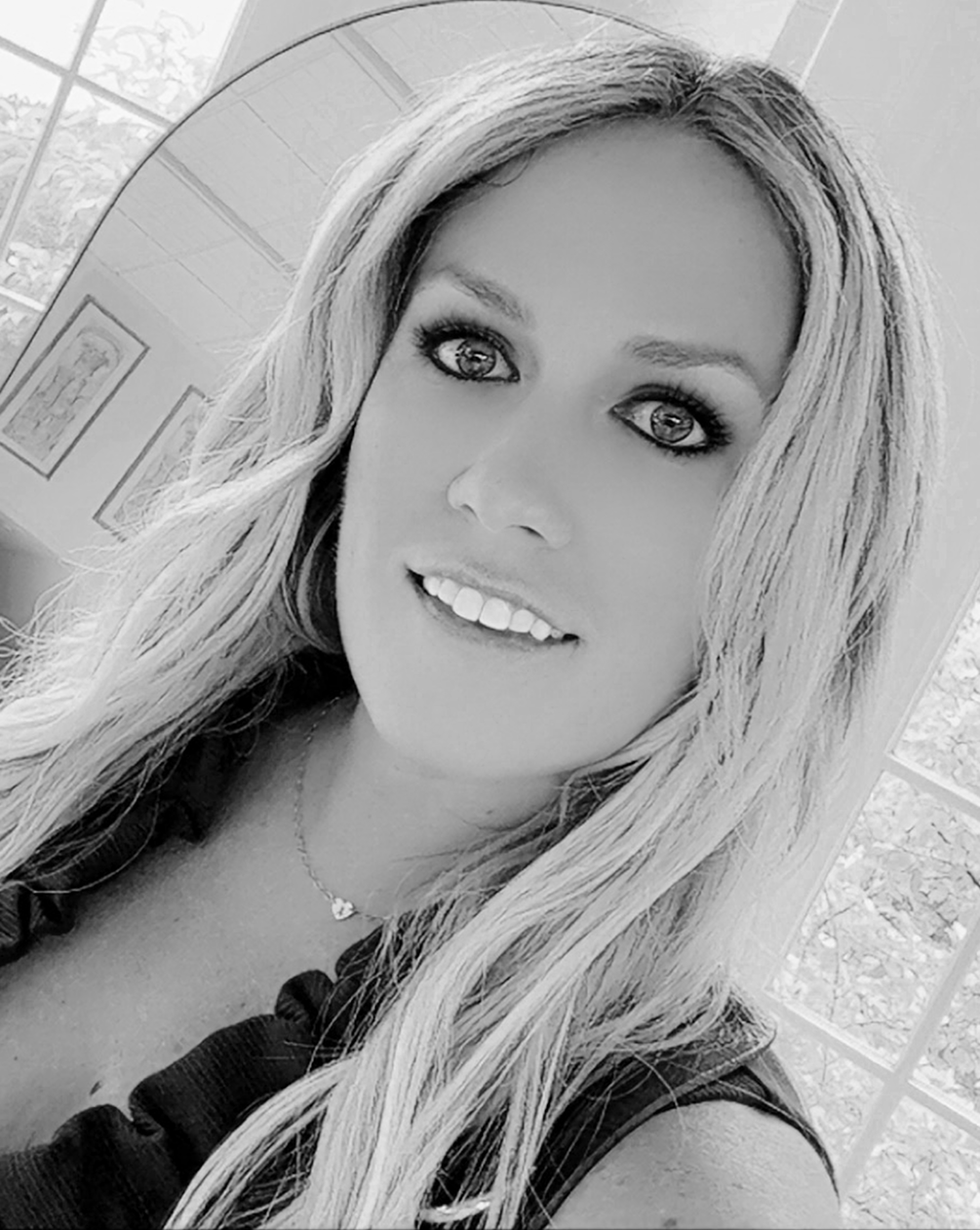 A woman with long wavy hair taking a selfie indoors, smiling at the camera. She is wearing a necklace and a ruffled top. The background includes a window with a view outside.