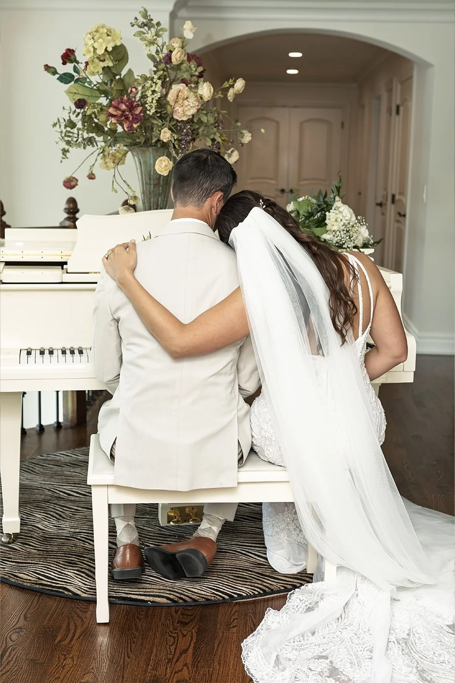 Bride and groom embracing in front of a white piano with a large floral arrangement on top, inside a decorated room.