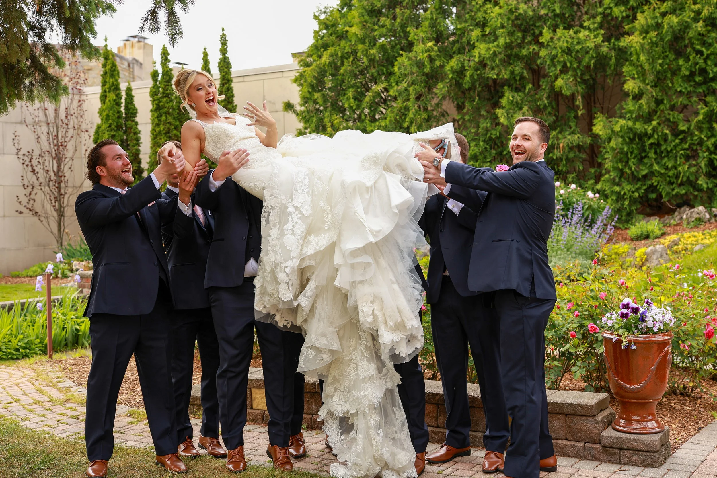 Group of groomsmen lifting a bride in her wedding dress outdoors in a garden.