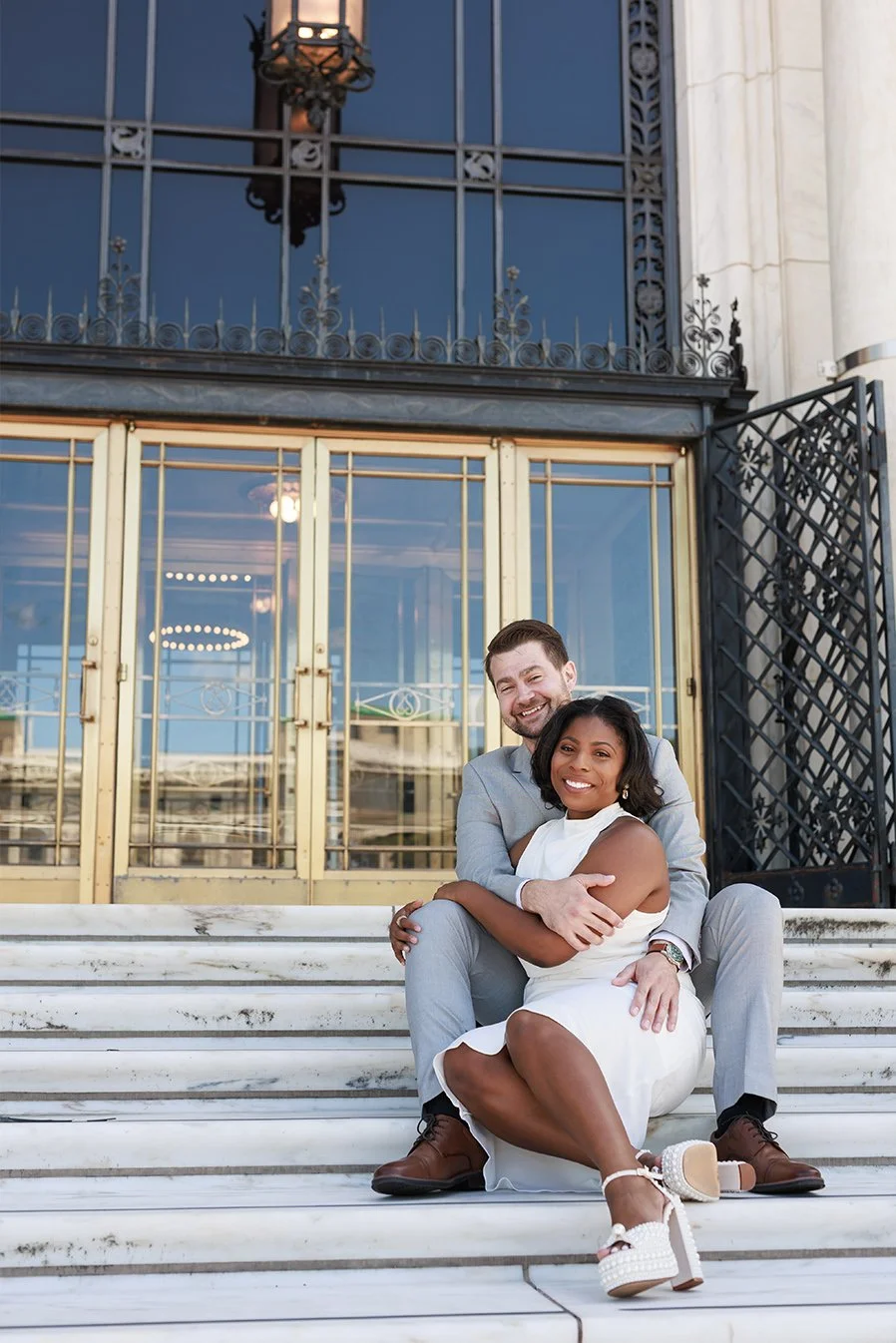 A happy couple sits on white marble stairs outside a building with large glass windows and metal decorative railings, smiling at the camera. Engagement photos at DIA in Detroit 