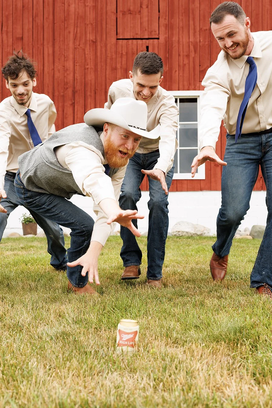 Four men wearing dress shirts and jeans gather outdoors on grass, watching a man in cowboy hat and vest reaching out toward a can with a Miller High Life label, with a red barn in the background. Fun wedding photo for groomsmen