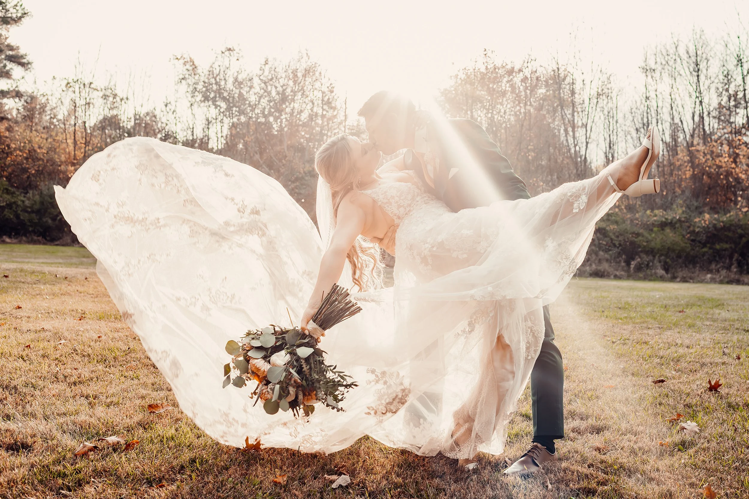 A bride and groom share a kiss outdoors in a sunlit park, with the bride being held horizontally by the groom. The bride is in a white lace wedding dress holding a bouquet, and the groom is in a dark suit. The sunlight creates a lens flare effect in 