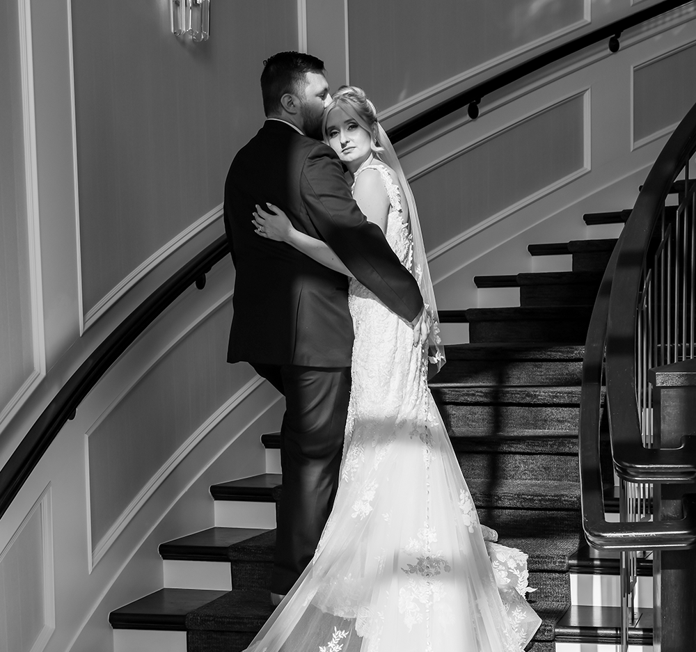 Black and white photo of a bride and groom embracing on a staircase, with the groom kissing the bride's forehead.