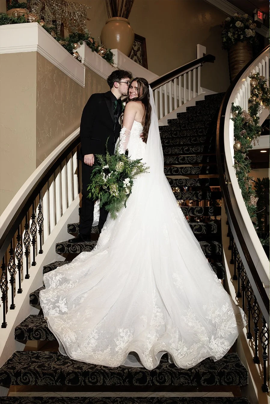 A newlywed couple on a staircase decorated for Christmas, with the bride in a white gown holding a floral bouquet and the groom in a black suit whispering to her.