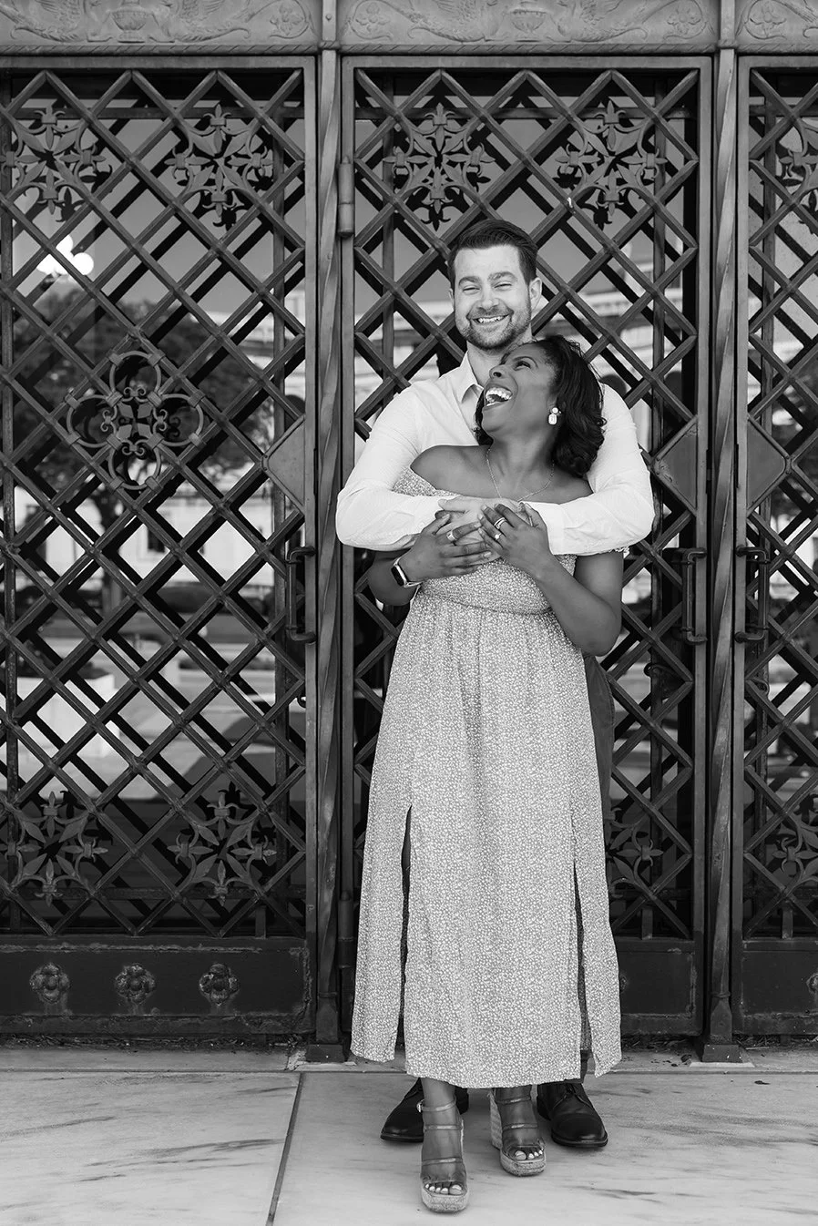 A happy couple laughing and hugging in front of a decorative metal gate, black and white engagement photo at the DIA Detroit.