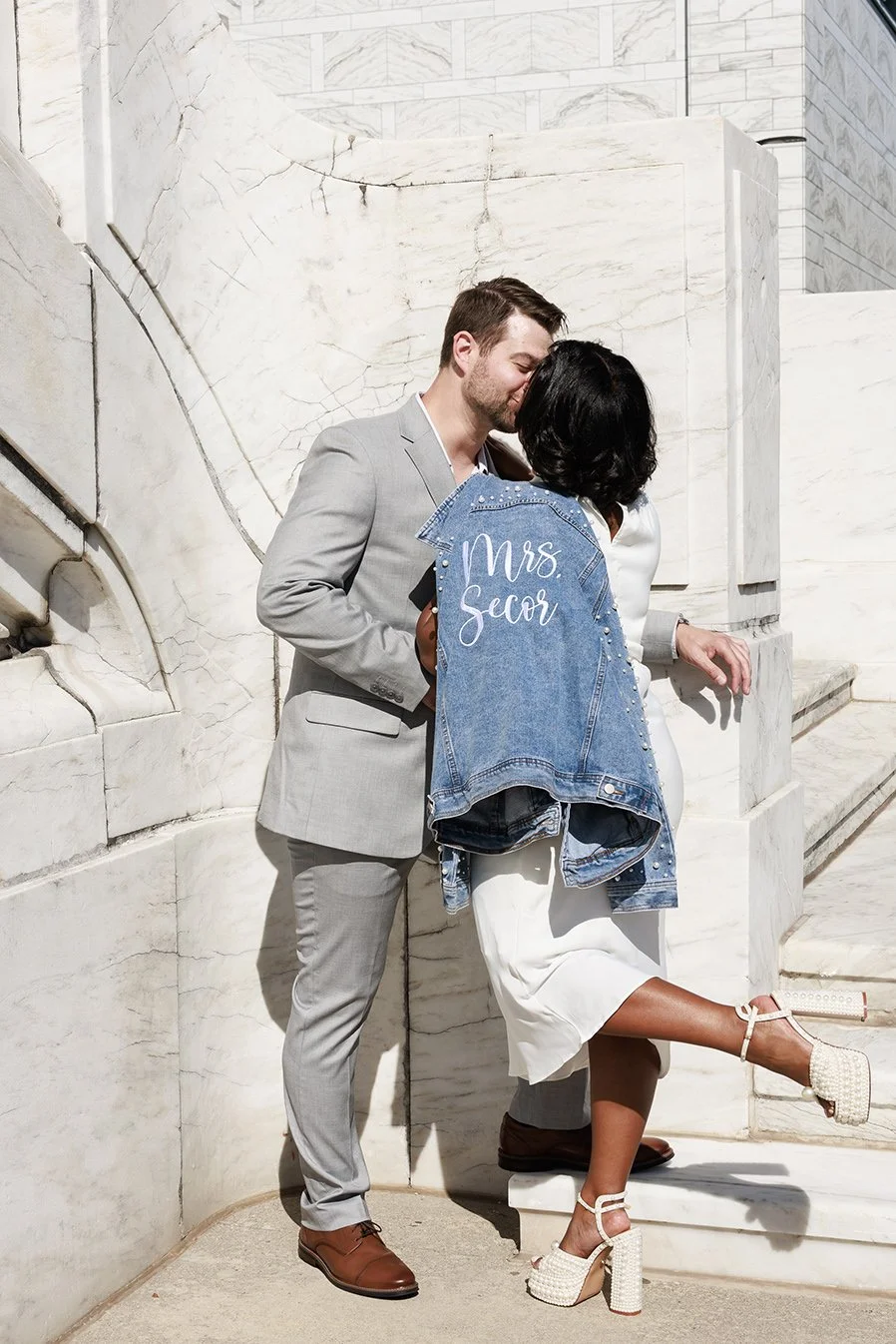 A couple sharing a kiss, standing on marble stairs near a white marble wall. The man is wearing a light gray suit and brown shoes. The woman is wearing white dress and high-heeled sandals, holding a denim jacket. Engagement photo at DIA in Detroit