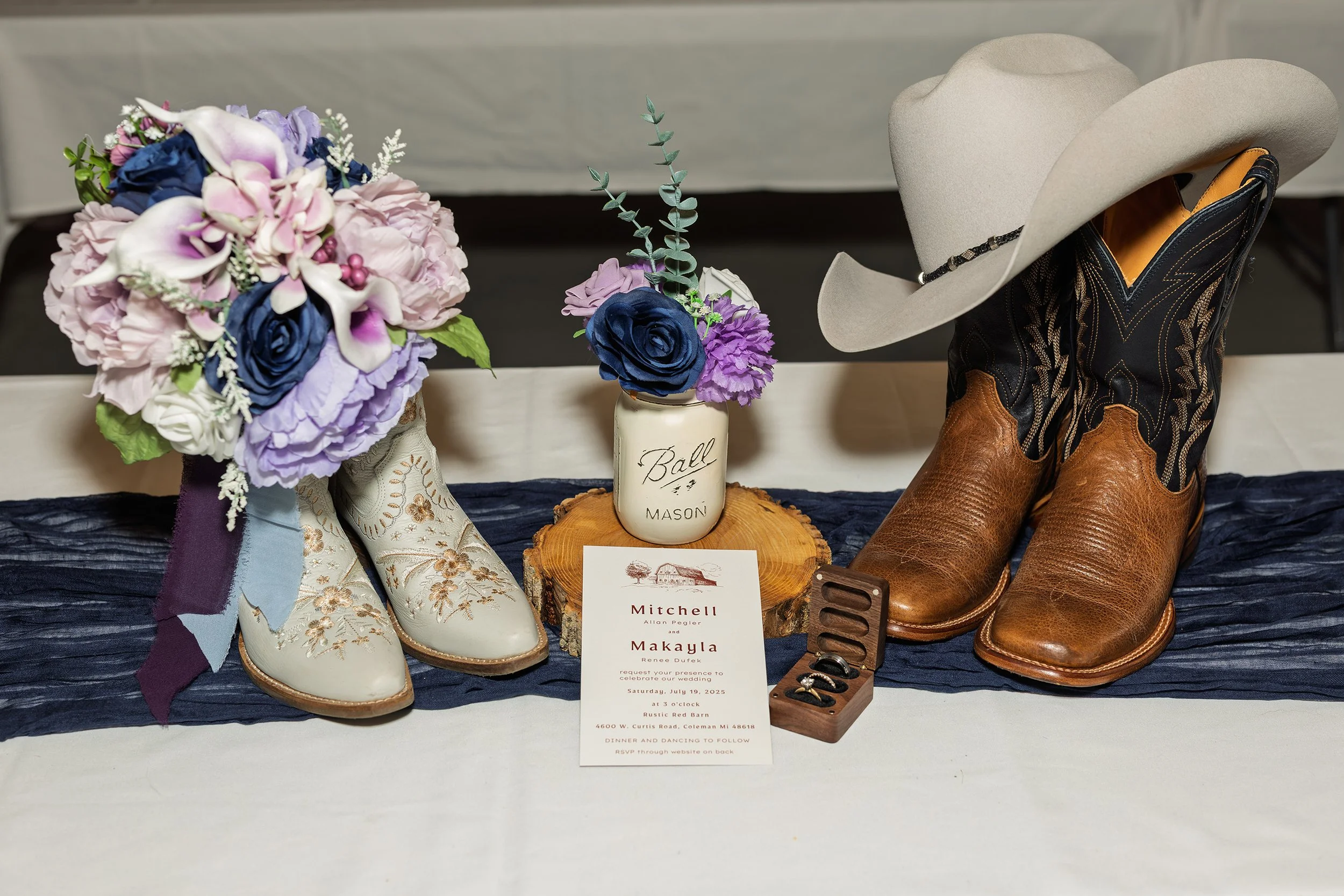 Wedding display with a bouquet of pink, purple, and blue flowers, a pair of white cowboy boots with floral embroidery, a small mason jar with flowers, a pair of brown cowboy boots with a wide hat placed on top, a ring box with wedding rings, and a we