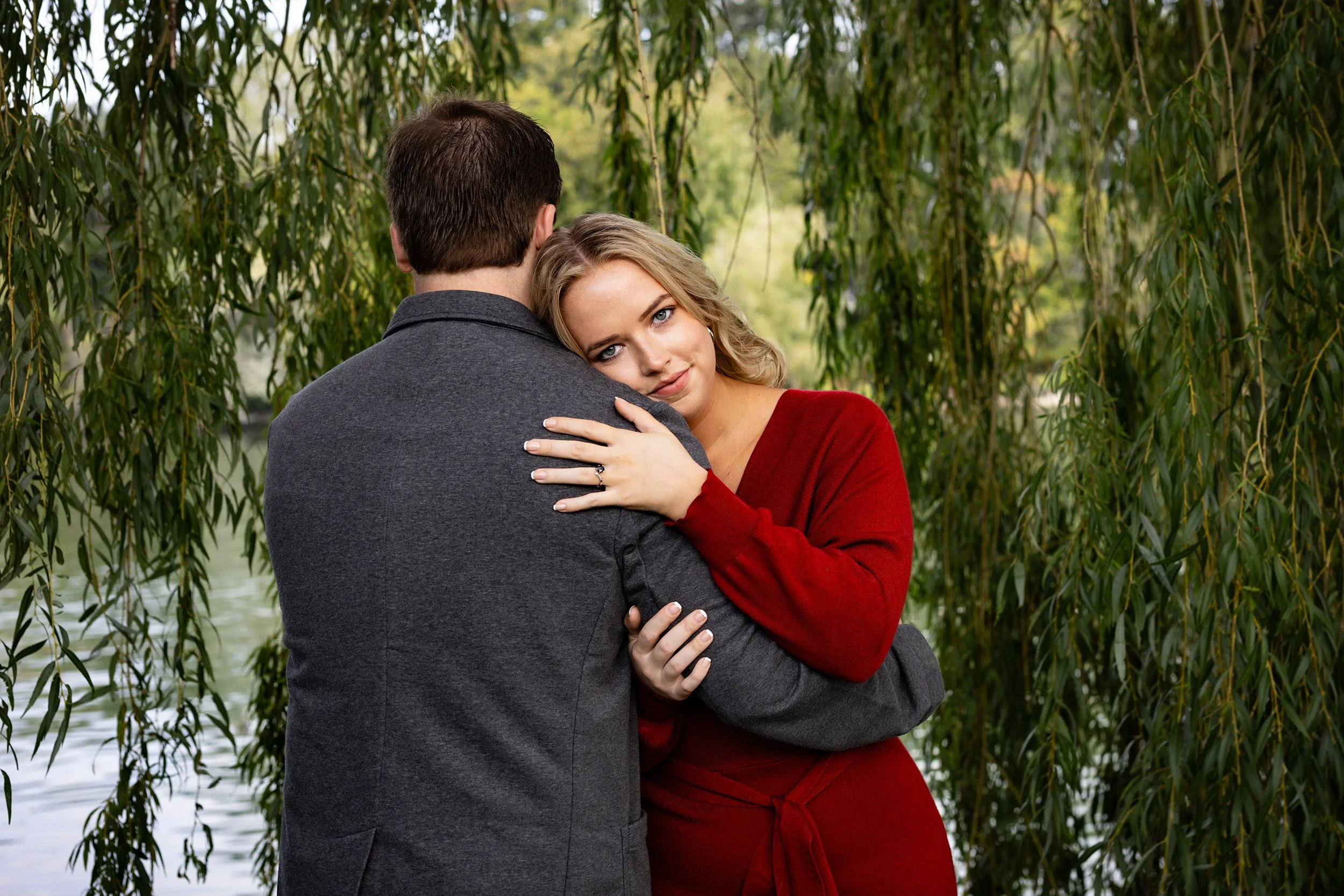 A young woman in a red dress leaning her head on a man's shoulder, embracing him outdoors near a body of water and surrounded by hanging willow tree branches, with green foliage in the background. Engagement photo