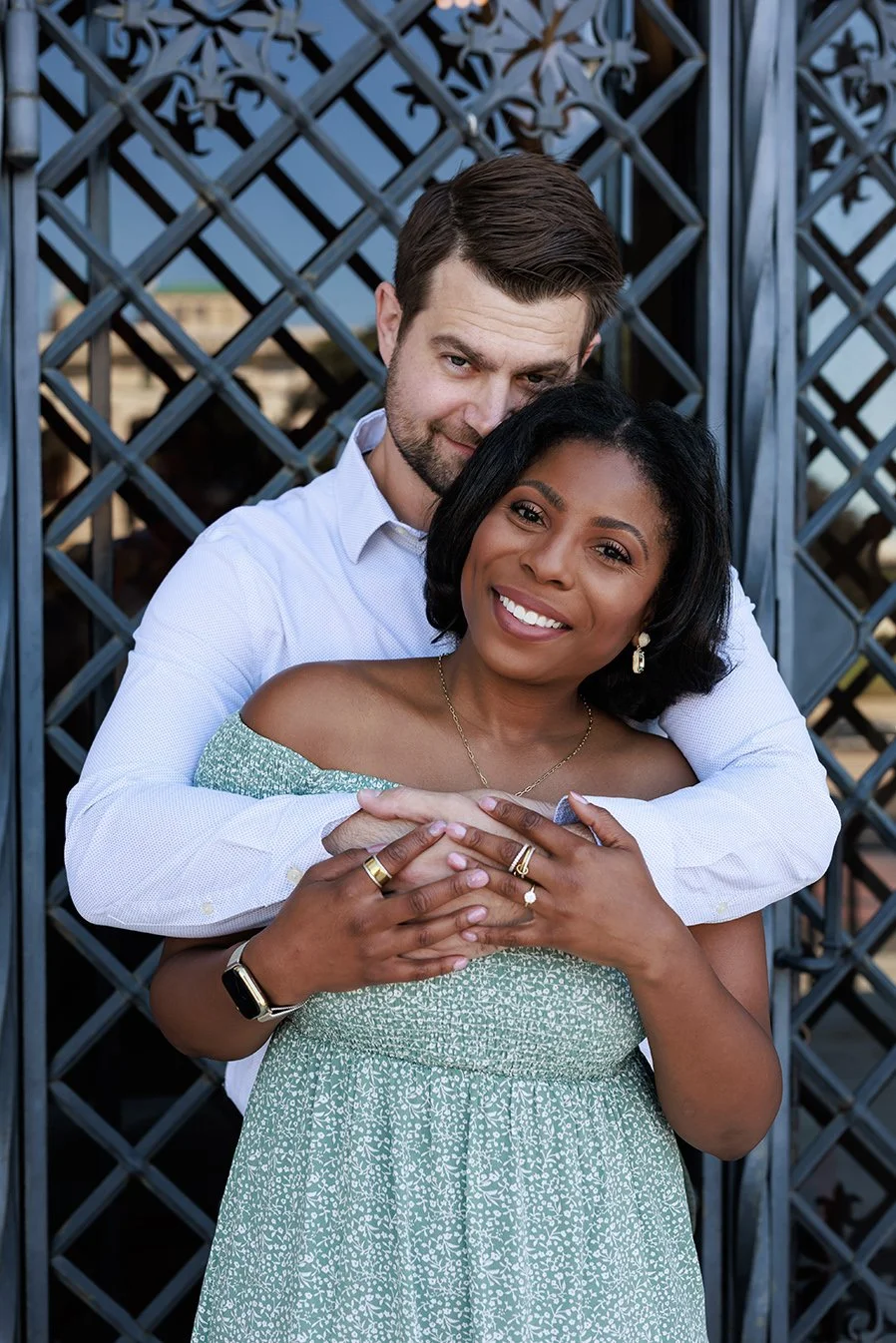 A man hugging a woman from behind, both smiling, standing outdoors in front of a decorative gate. Engagement photo at DIA in detroit