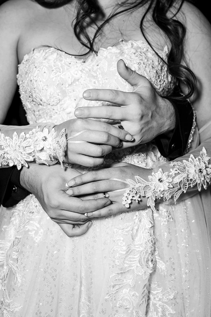 Black and white photograph of a bride and groom holding each other's hands. The bride is wearing a lace wedding dress with floral embroidery and lace gloves. The groom's hand, with a wedding ring, is resting over the bride's waist. The focus is on th