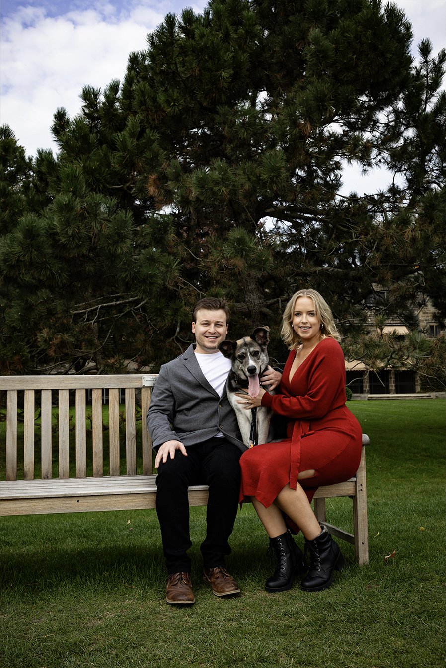 A smiling man and woman sitting on a wooden bench outdoors with a dog between them. The woman is wearing a red dress and black boots, and the man is in a gray blazer and black pants. There is a large tree with green foliage in the background and a grassy area.
