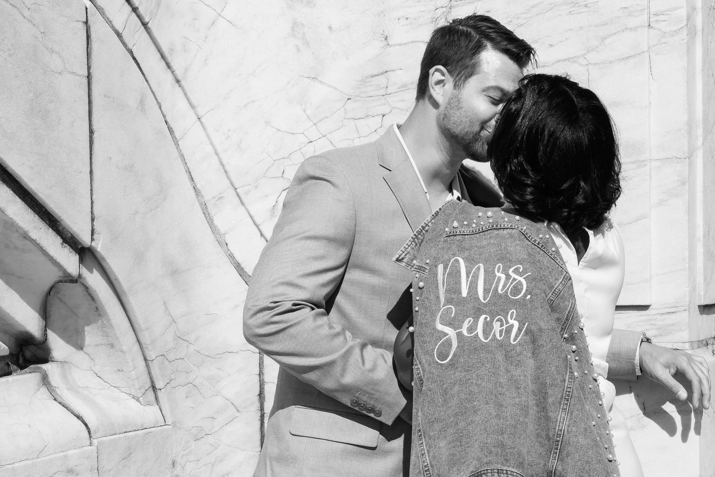 A black and white photo of a man and woman kissing. The woman is holding a denim jacket with "Mrs. Secor" written on it. The man is wearing a suit, and they are standing against a marble wall.