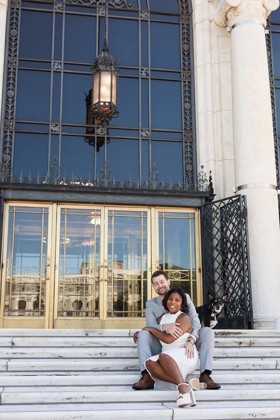 A happy couple sitting on white marble stairs outside a historic building with large glass doors and intricate ironwork, with a black and white dog sitting behind them. Engagement photo at DIA in Detrit