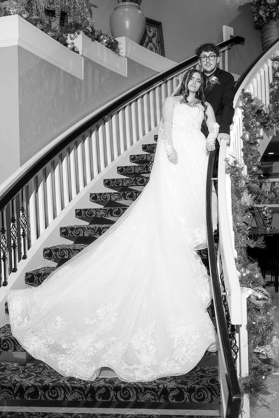 Black and white photo of a bride and groom standing on a staircase decorated with Christmas garland. The bride wears a long white wedding gown with lace gloves, and the groom in a dark suit with glasses.