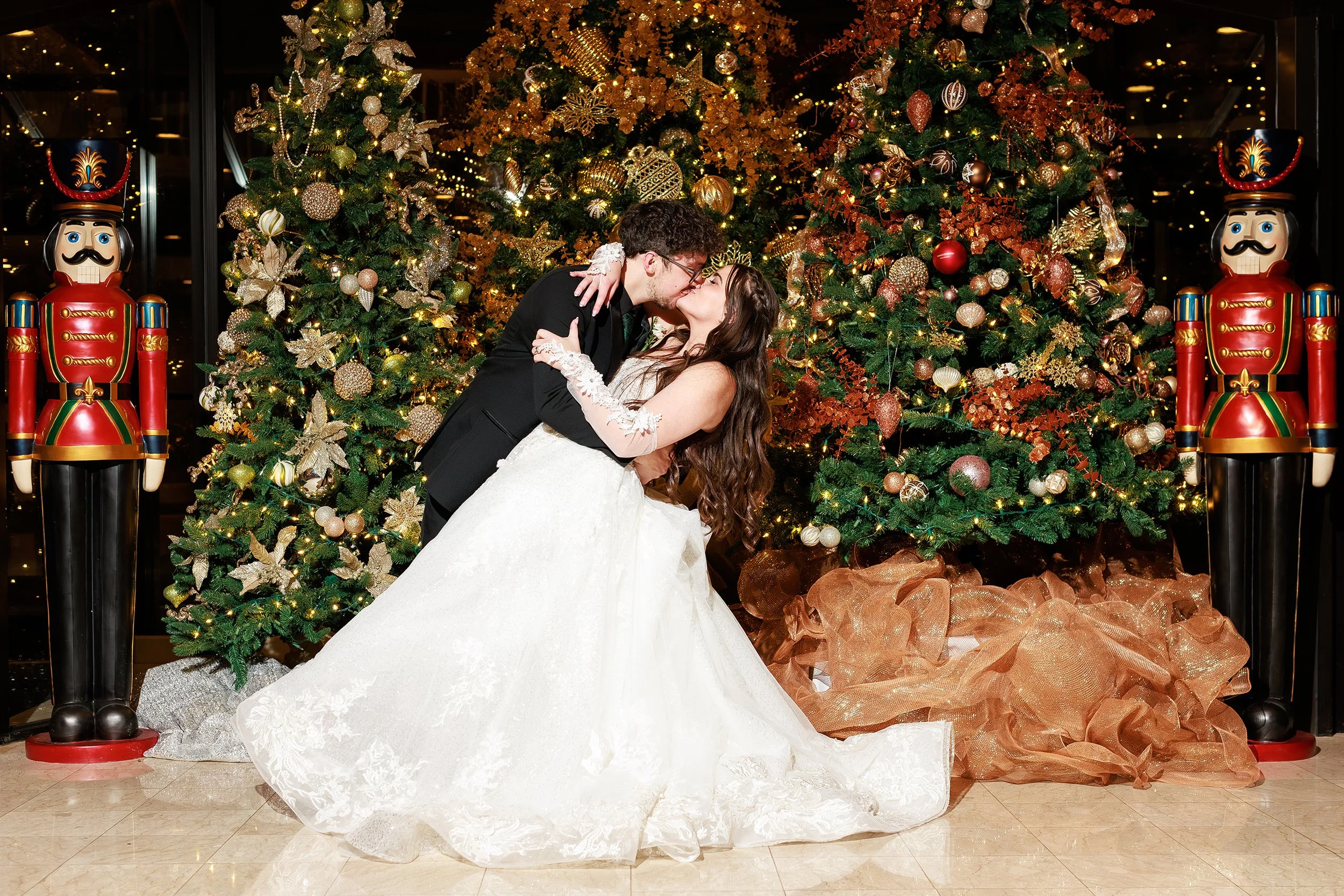 A newlywed couple sharing a kiss in front of decorated Christmas trees with ornaments and lights, flanked by two large nutcracker figurines.