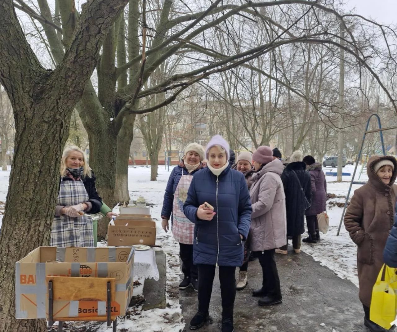 A group of people, mostly women, are gathered outdoors in a snowy park. Some are standing in line, while others are talking or standing near tables with boxes. The scene appears to be a community event or outdoor market during winter.