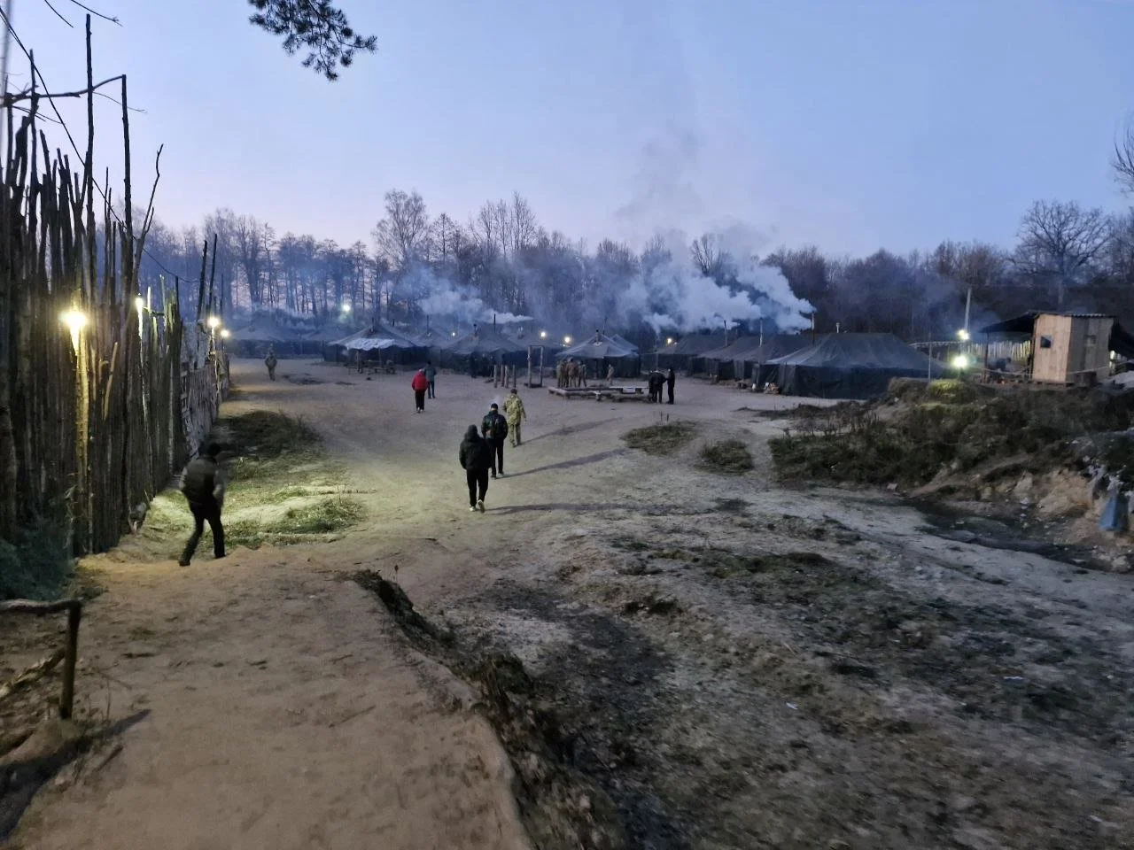 A dirt pathway leading to a village with tents and smoke rising in the background, with several people walking and lights illuminating the scene during dusk.