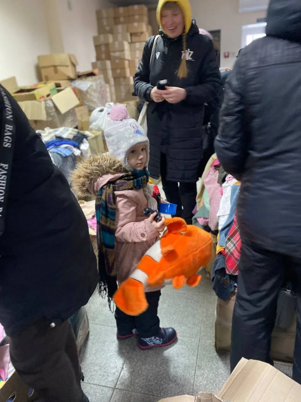 A young girl with a fur-lined hooded coat, colorful scarf, and boots, holding a plush fish in a crowded room with stacks of boxes and clothing; a woman with a yellow hat and black jacket is smiling nearby.