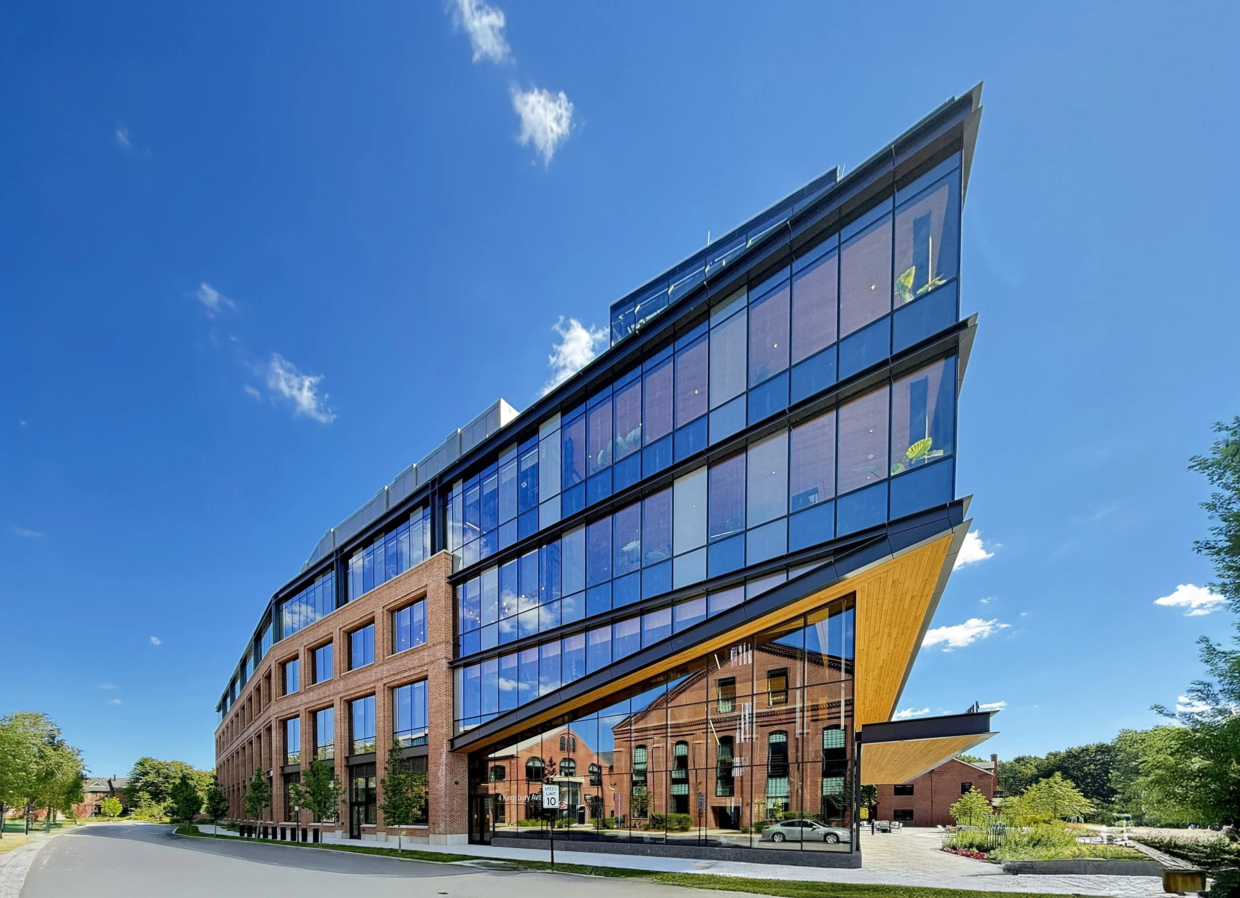 Modern multi-story building with glass façade and brick accents, situated on a tree-lined street on a sunny day. Massachusetts, new construction, life science, watertown, architecture