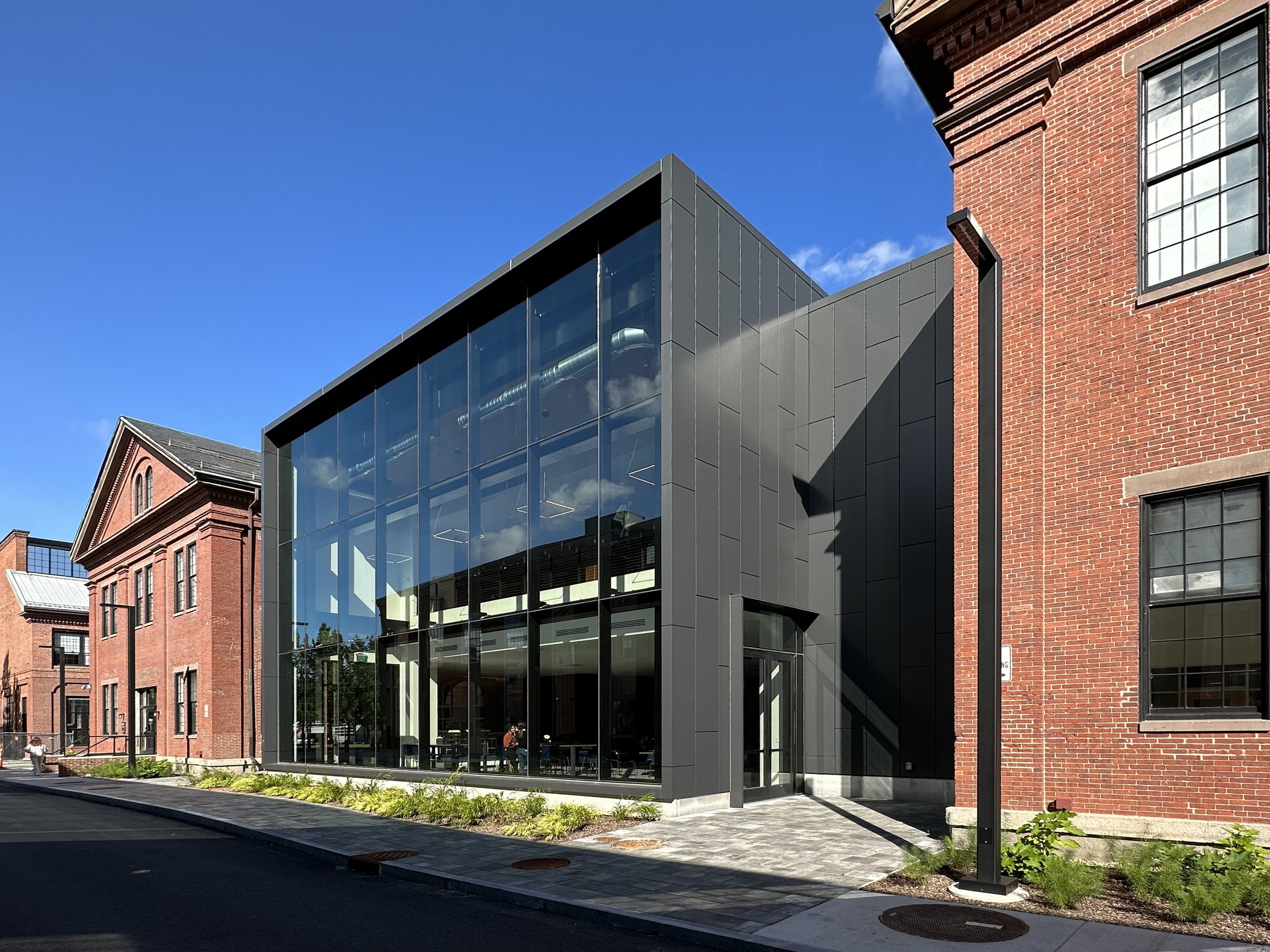 Modern glass building with dark gray exterior panels adjacent to red brick buildings, under a blue sky. Massachusetts, new construction, life science, watertown, architecture