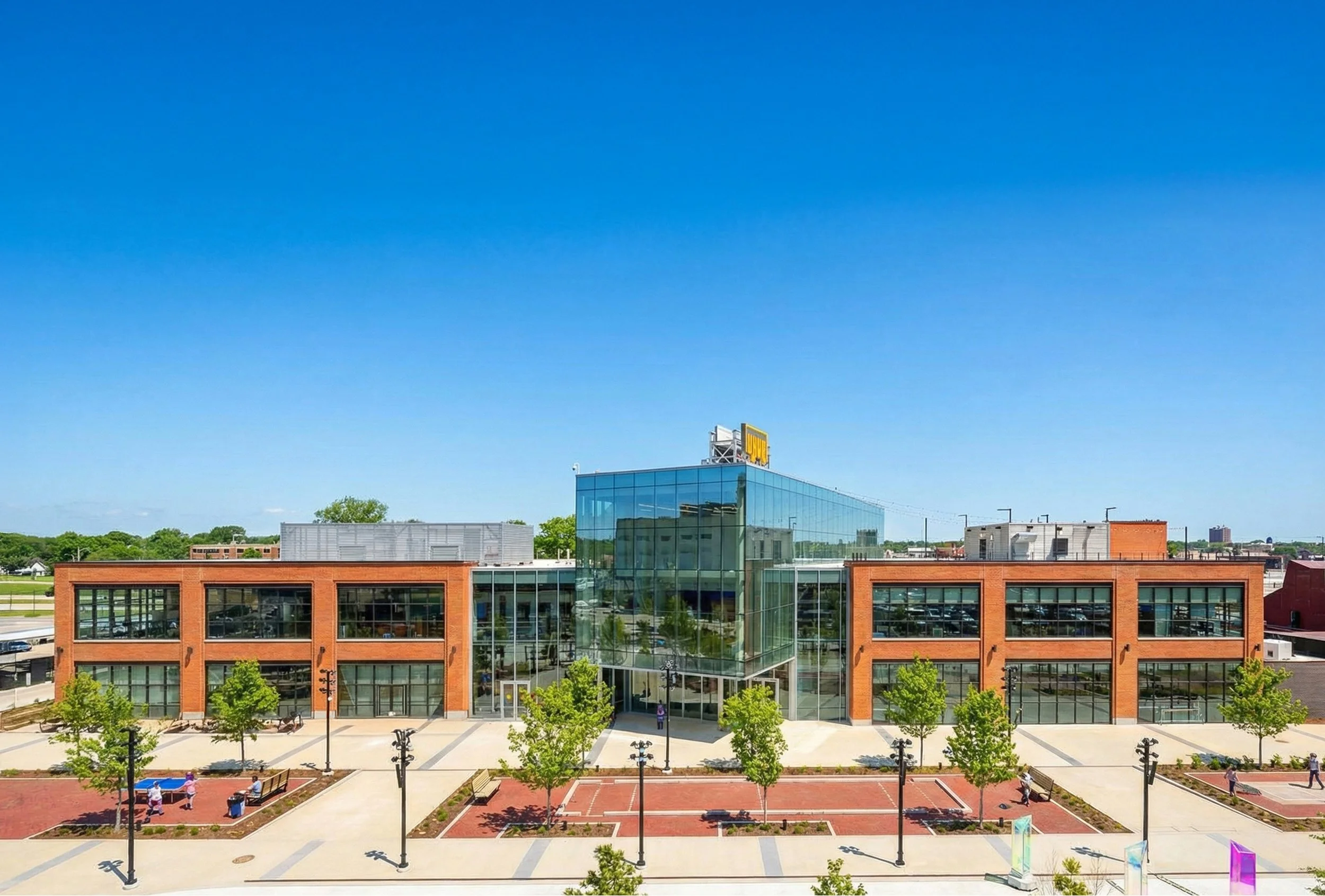 Modern building with red brick and glass facade, surrounded by trees, benches, and open plaza, under a clear blue sky. Architecture, new construction, green bay, packers, lambeau