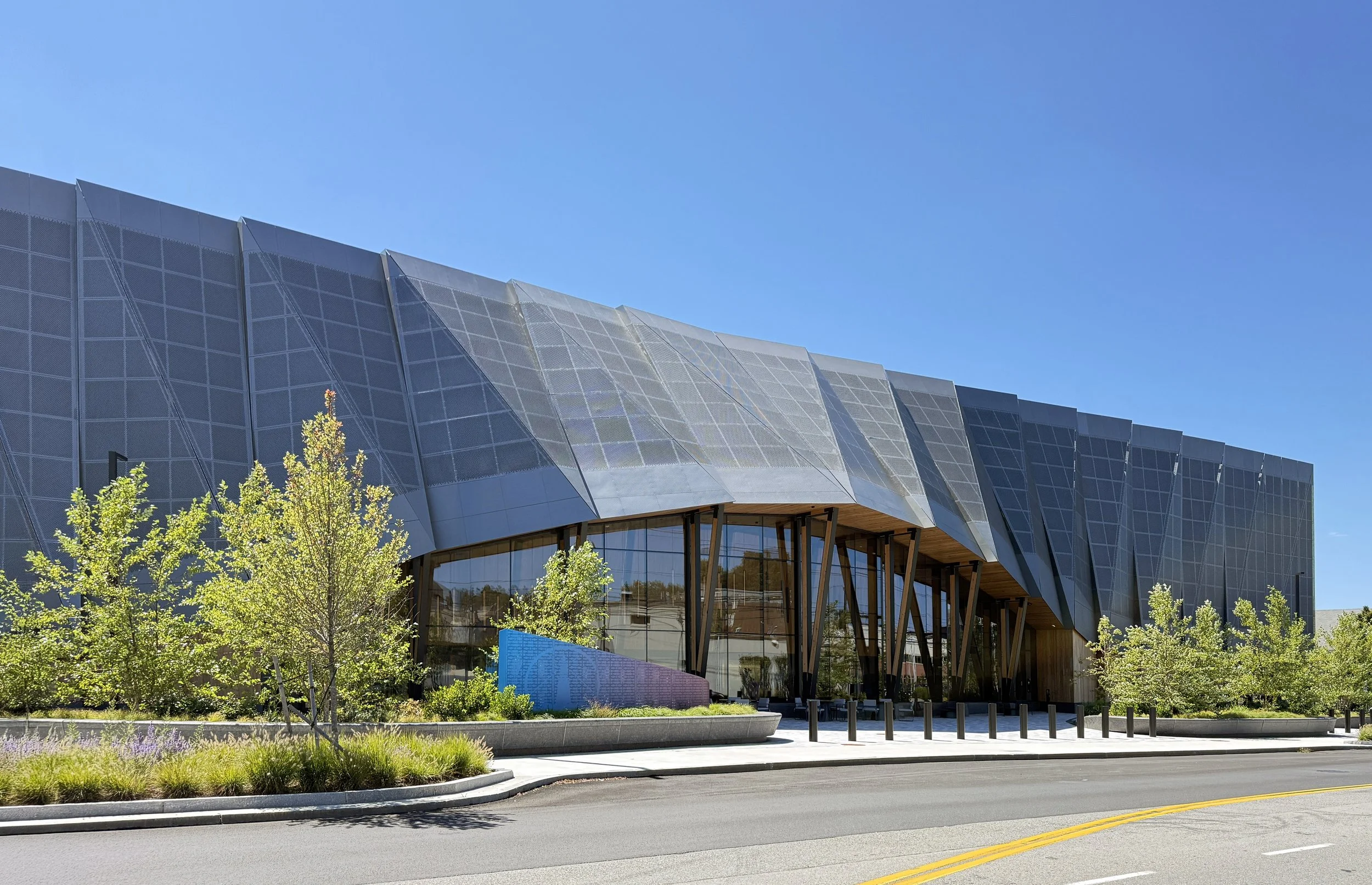 Modern building with angular metallic panels, glass entrance, and surrounding greenery under a clear blue sky. Massachusetts, new construction, transit up, watertown, architecture