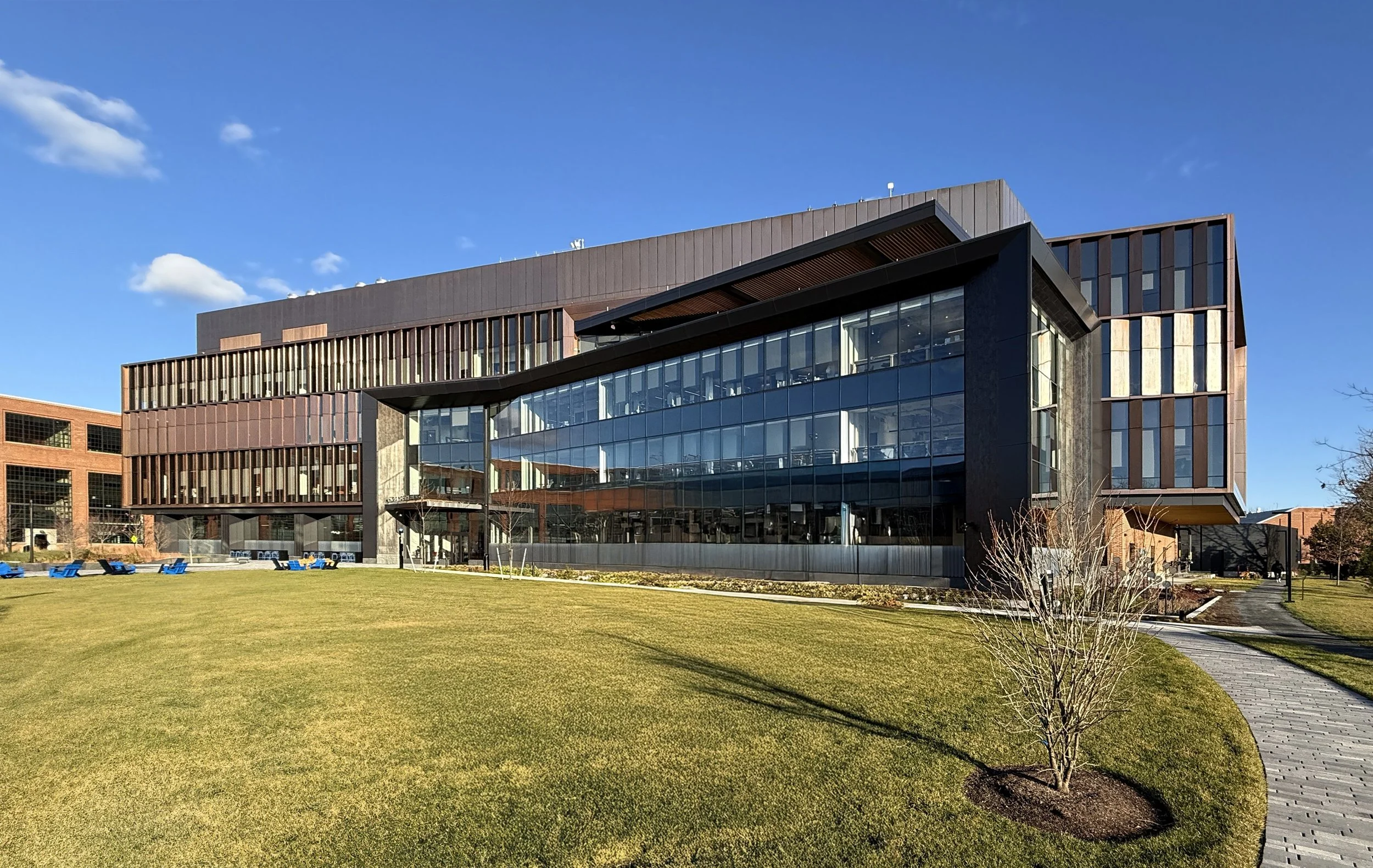 Modern office building with glass windows, surrounded by a well-kept lawn and walking path, under a clear blue sky. Massachusetts, new construction, life science, watertown, architecture