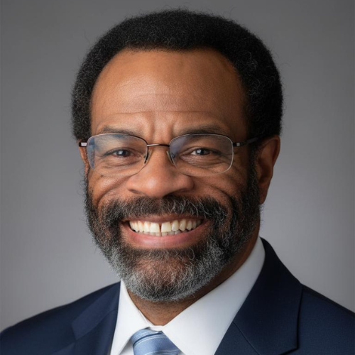 Headshot of an African American man with glasses, a beard, and a mustache, smiling and wearing a dark suit and striped tie.