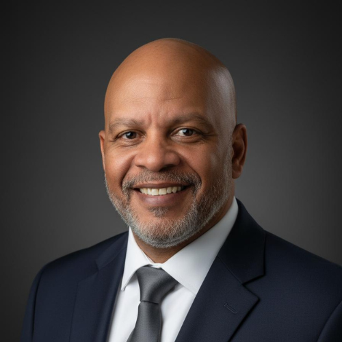 Close-up portrait of a smiling bald African American man in a dark suit, white shirt, and gray tie, against a dark gray background.