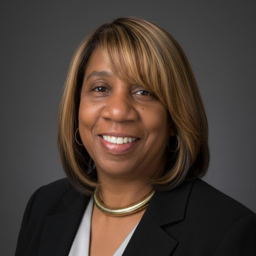 Professional portrait of an African American woman with shoulder-length hair, wearing a black blazer, white top, gold necklace, and small hoop earrings, smiling against a dark gray background.
