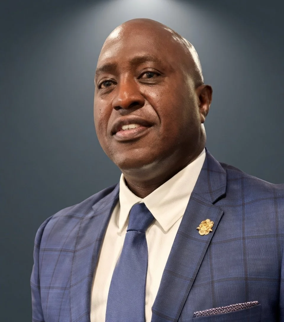 A professional man wearing a blue checkered suit, white shirt, and a blue tie, with a gold pin on his lapel, posing against a dark background.