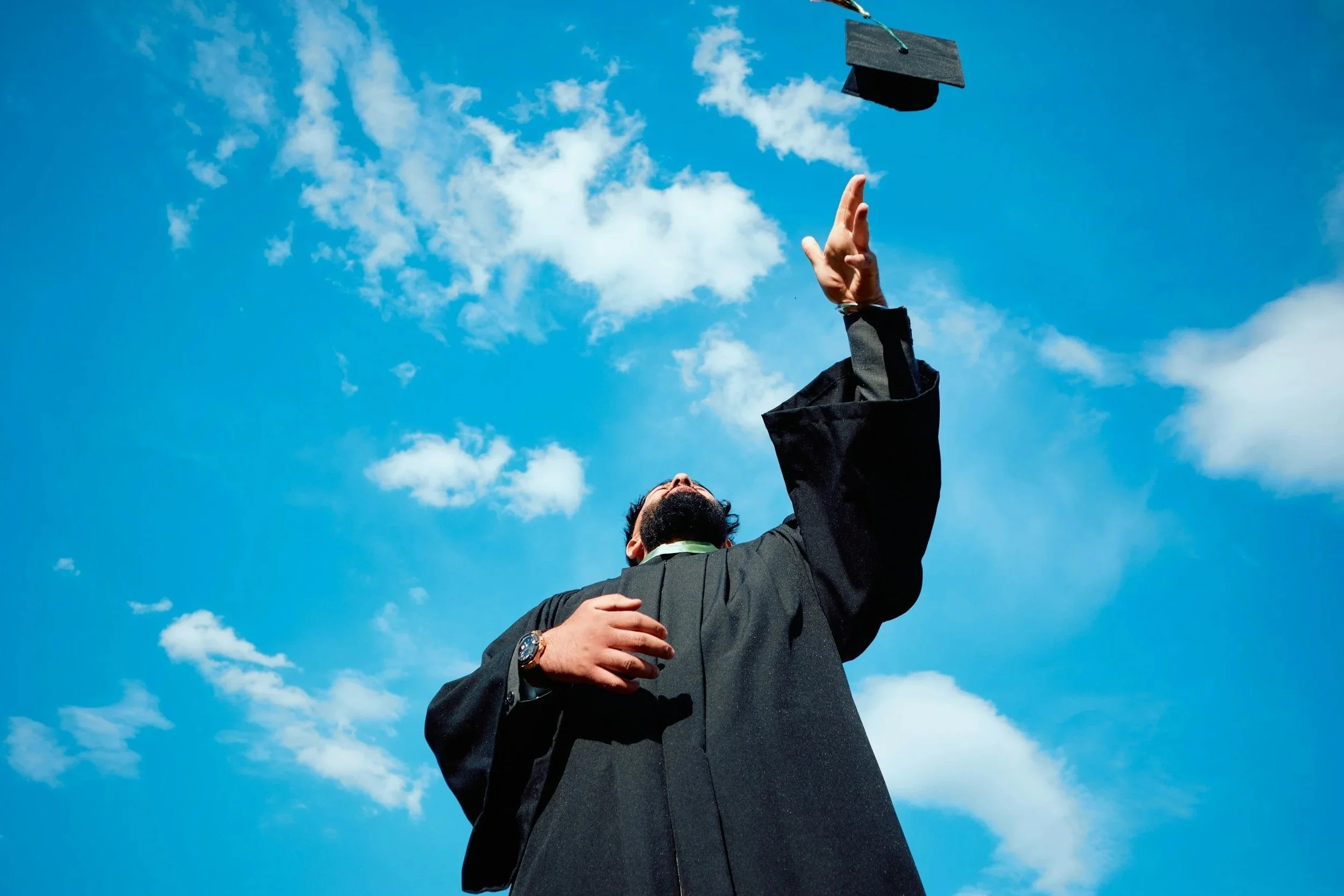 A person in a black suit throwing a graduation cap into the air outdoors against a blue sky with scattered clouds.