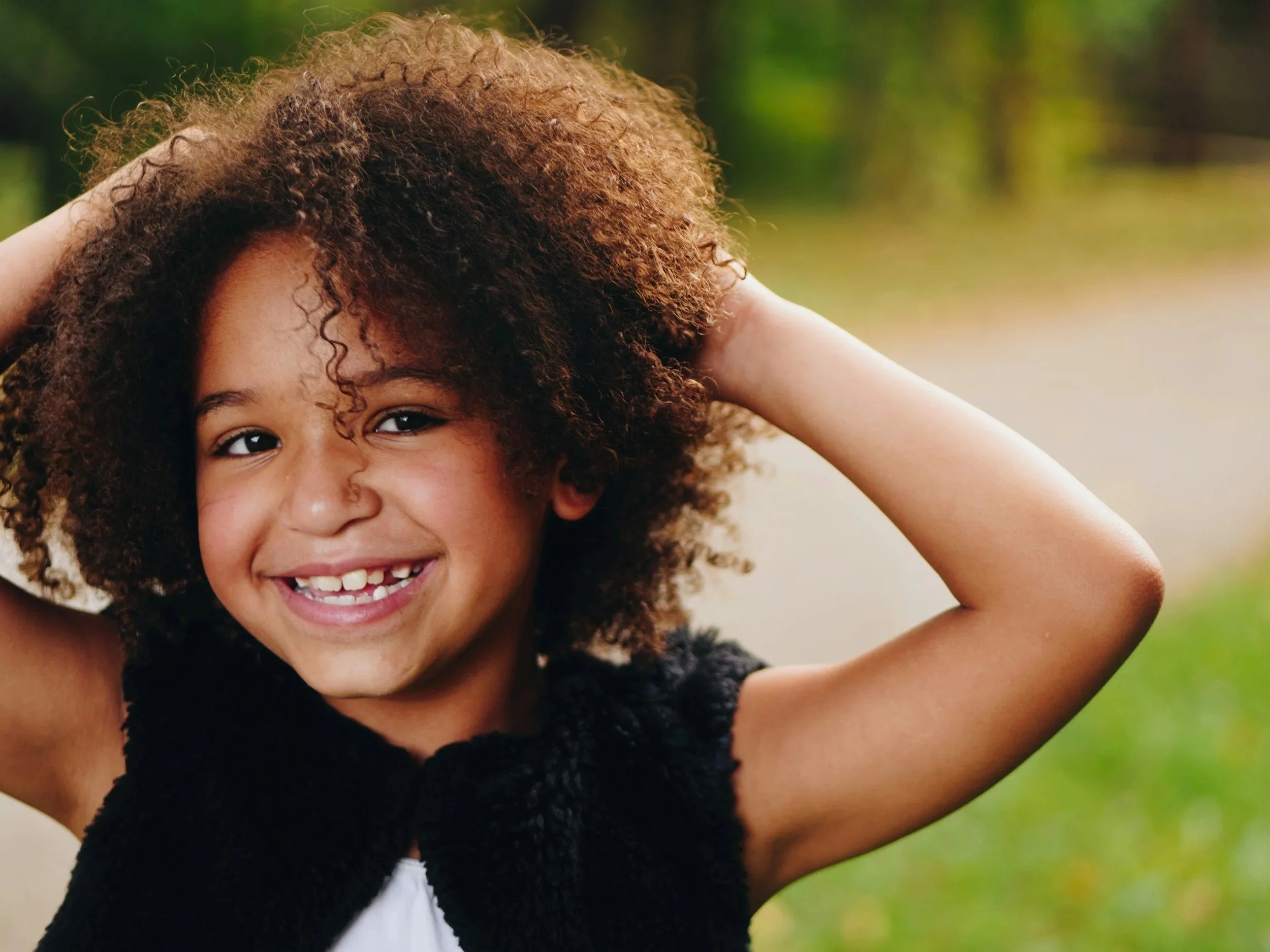 Close-up of a young girl smiling with curly hair outdoors.